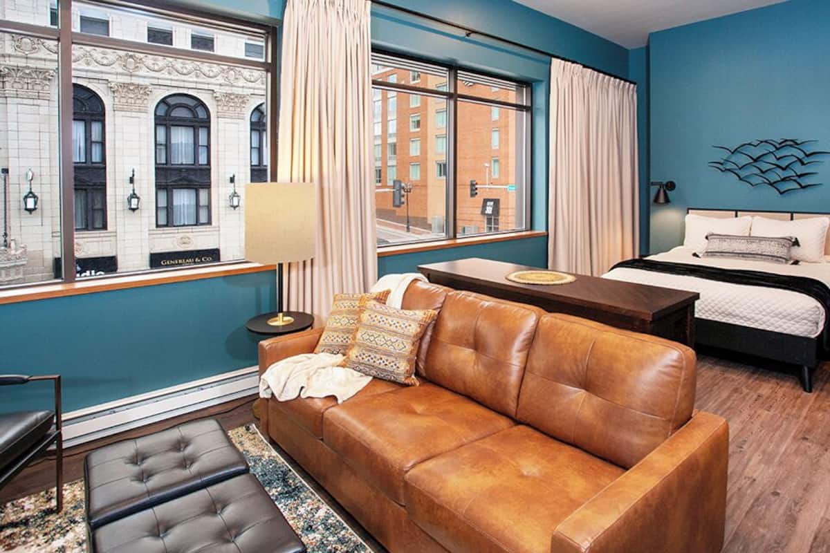 A modern hotel room with a large window, a brown leather sofa, a bed with a white headboard, and a small table.