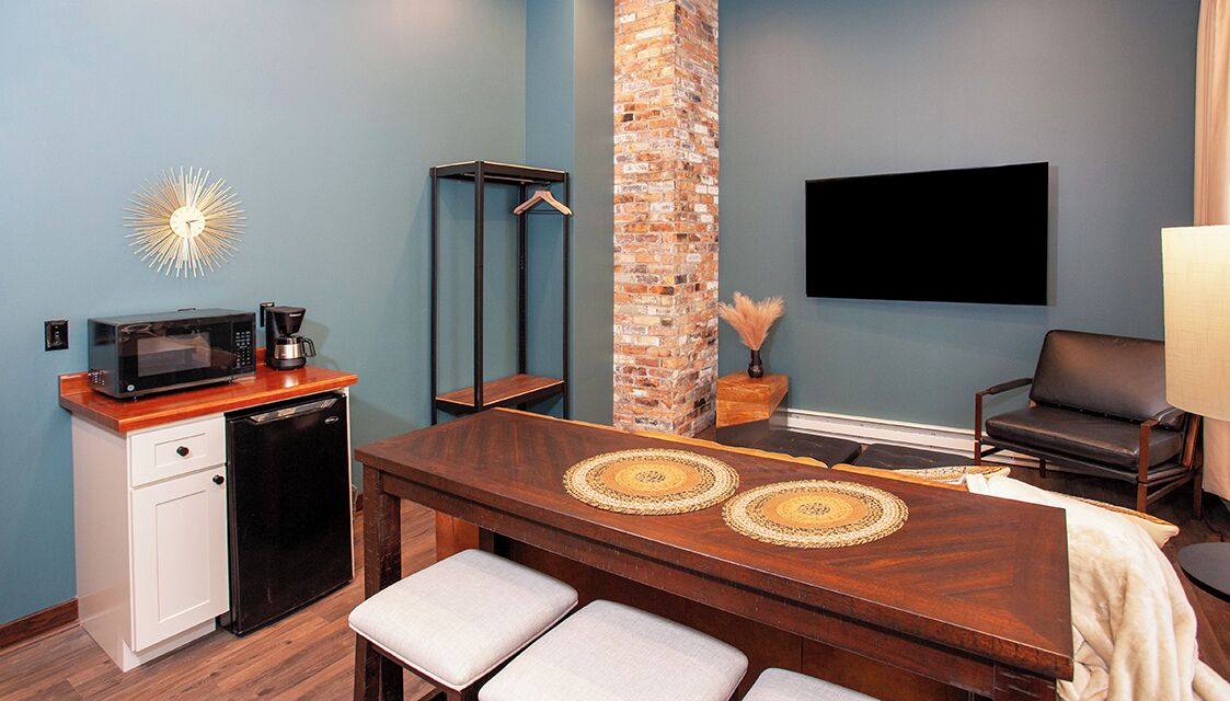 A compact room with a wooden dining table, a small kitchenette with a microwave and coffee maker, a brick accent wall, a hanging coat rack, and a flat-screen TV mounted on the wall.