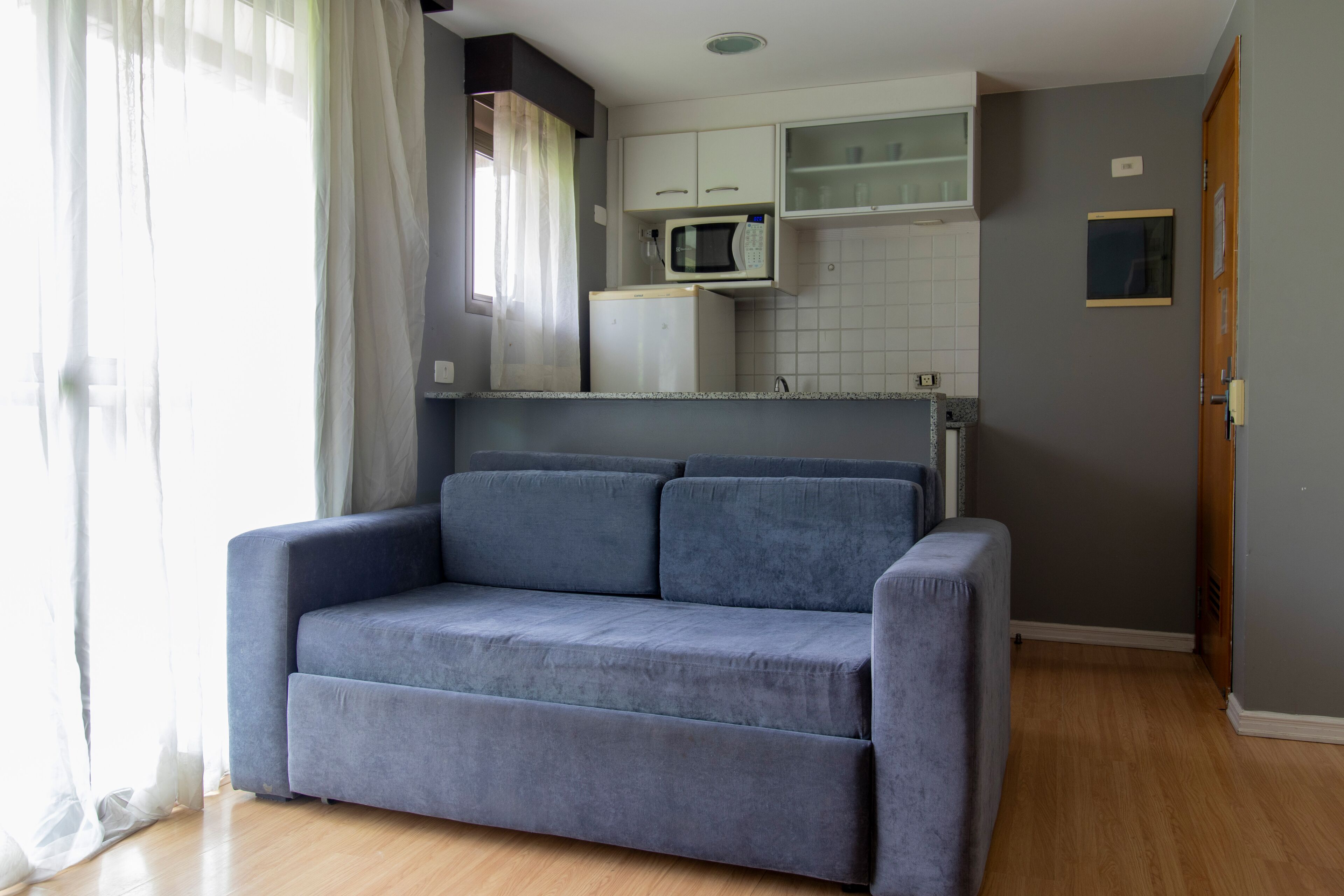 Ein modernes Wohnzimmer mit grauem Sofa, einer Küchenzeile inklusive Mikrowelle und Holzboden.