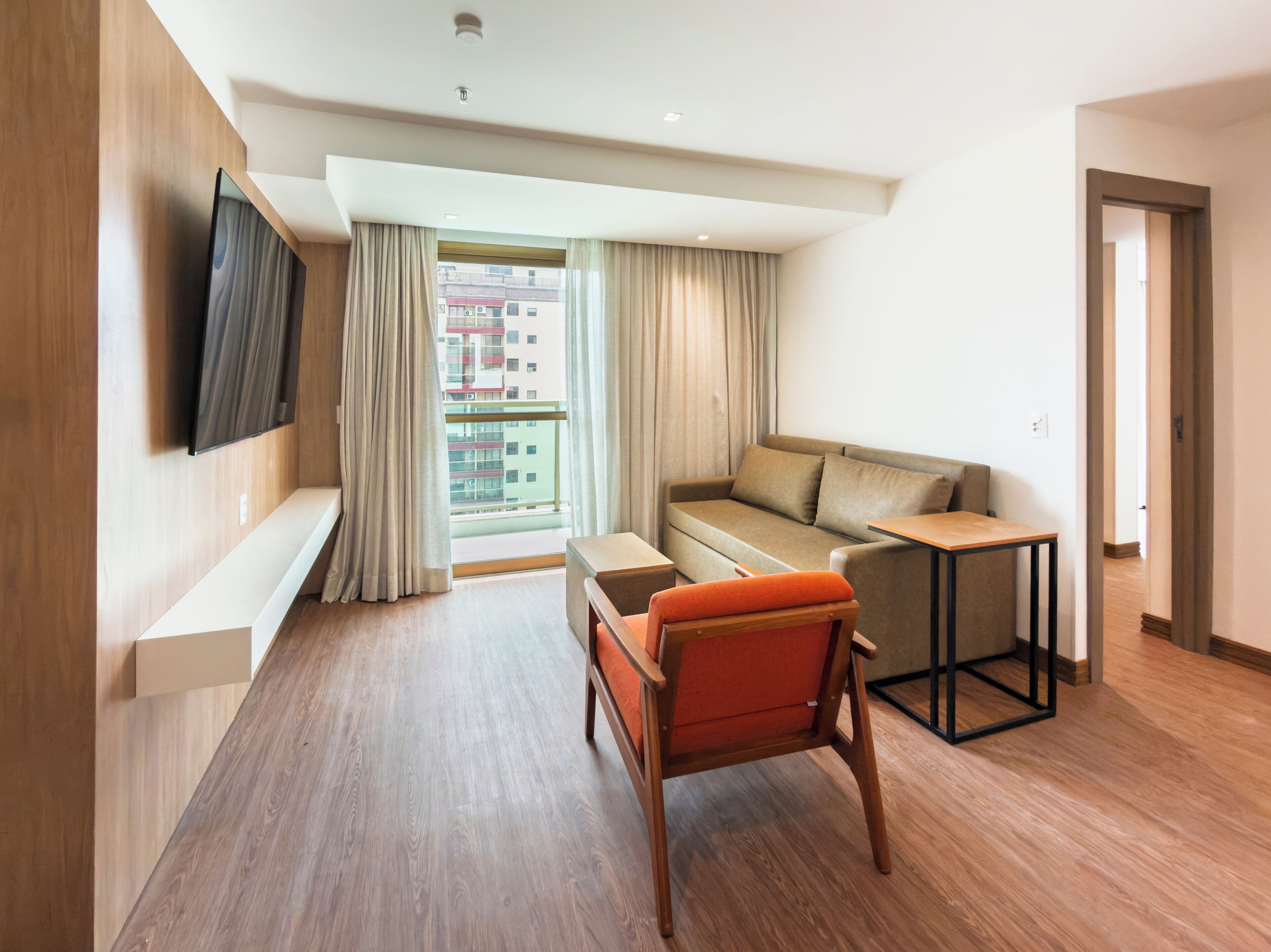 A modern hotel room with a wooden floor, a sofa, an orange chair, a small table, and a TV mounted on the wall.