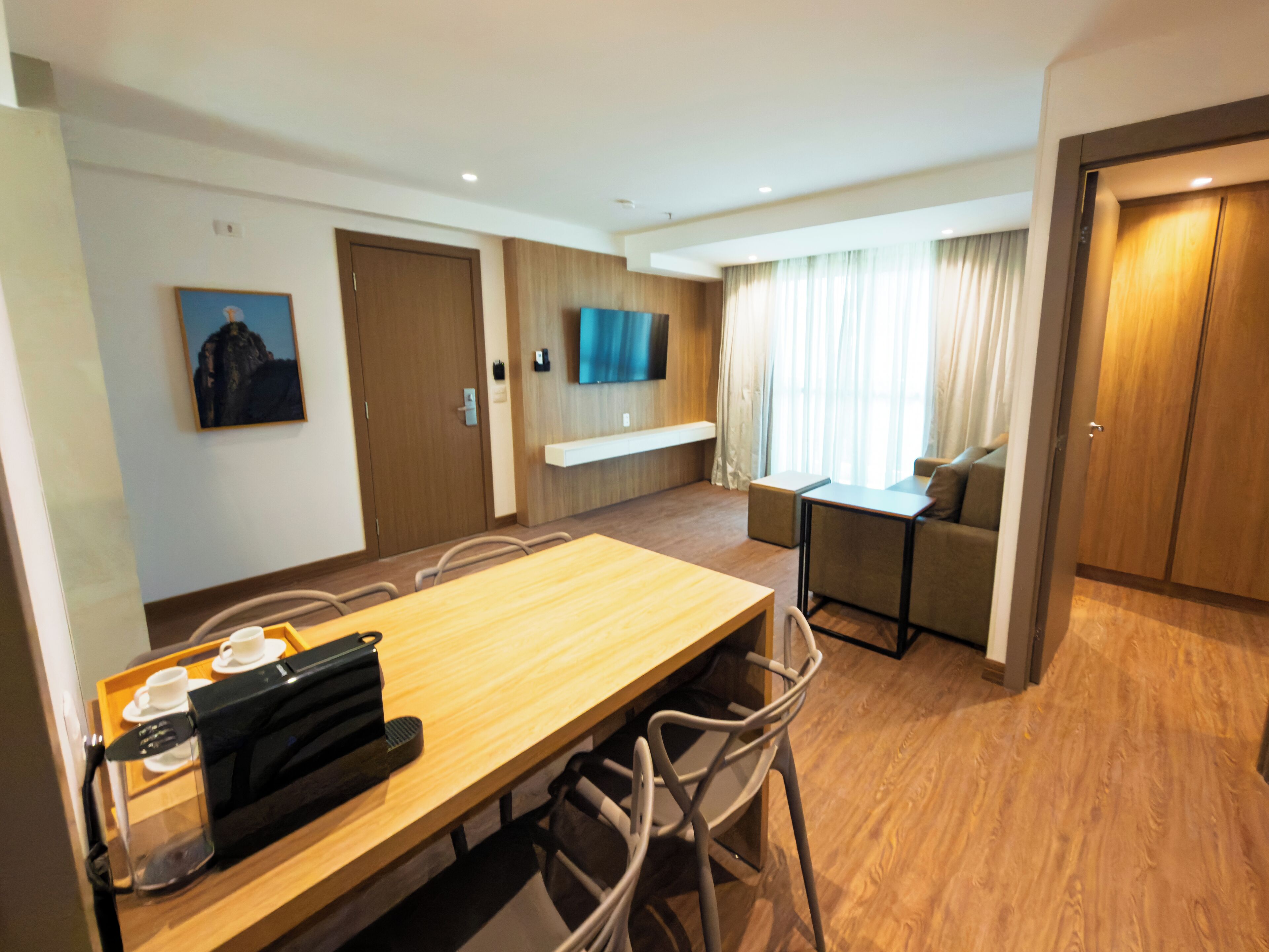 A hotel room with a wooden dining table, chairs, a coffee machine, a TV mounted on the wall, a sofa, and a window with curtains.
