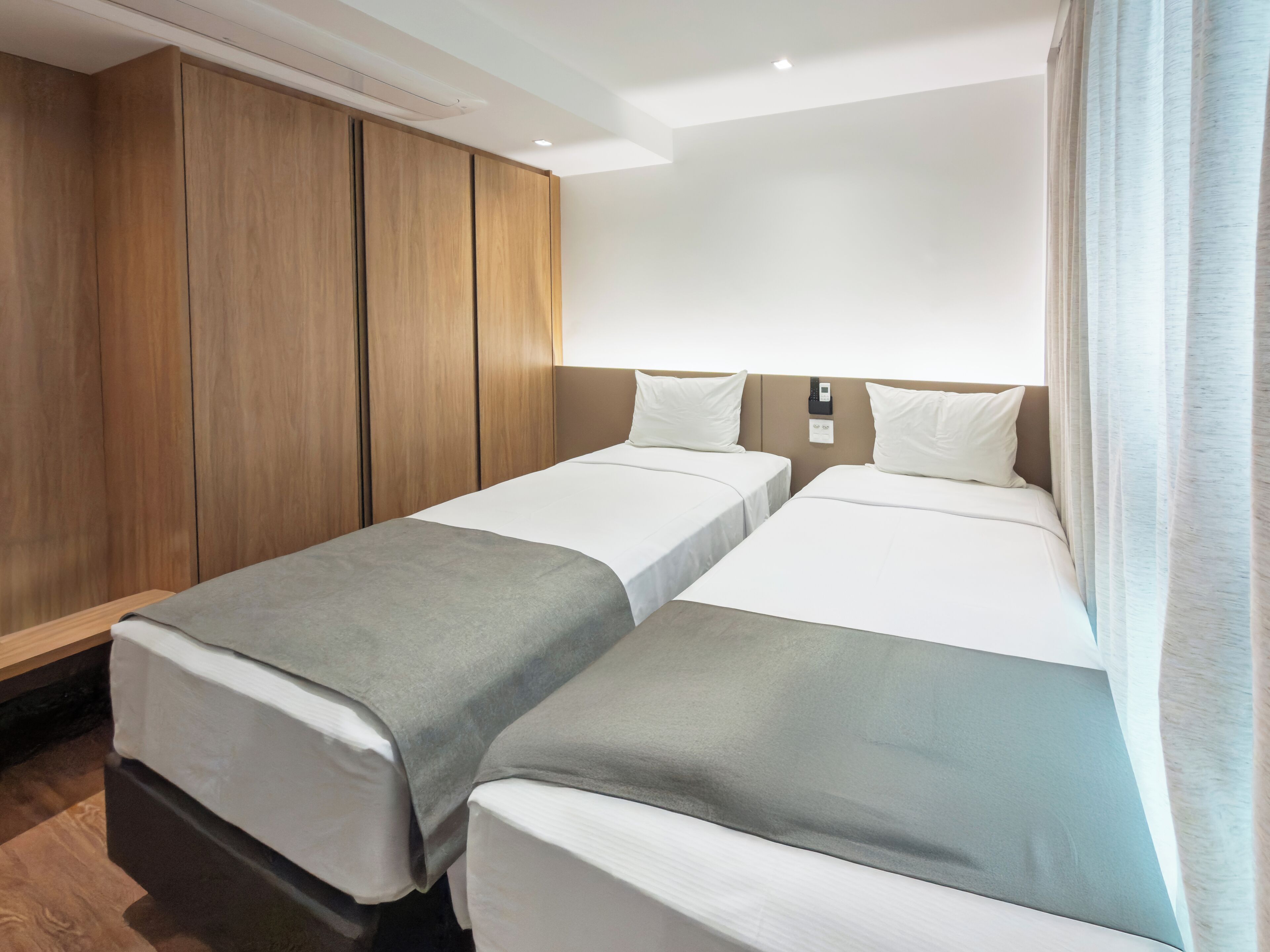 Una habitación de hotel con dos camas individuales, un armario de madera, una mesita de noche y una ventana con cortinas.