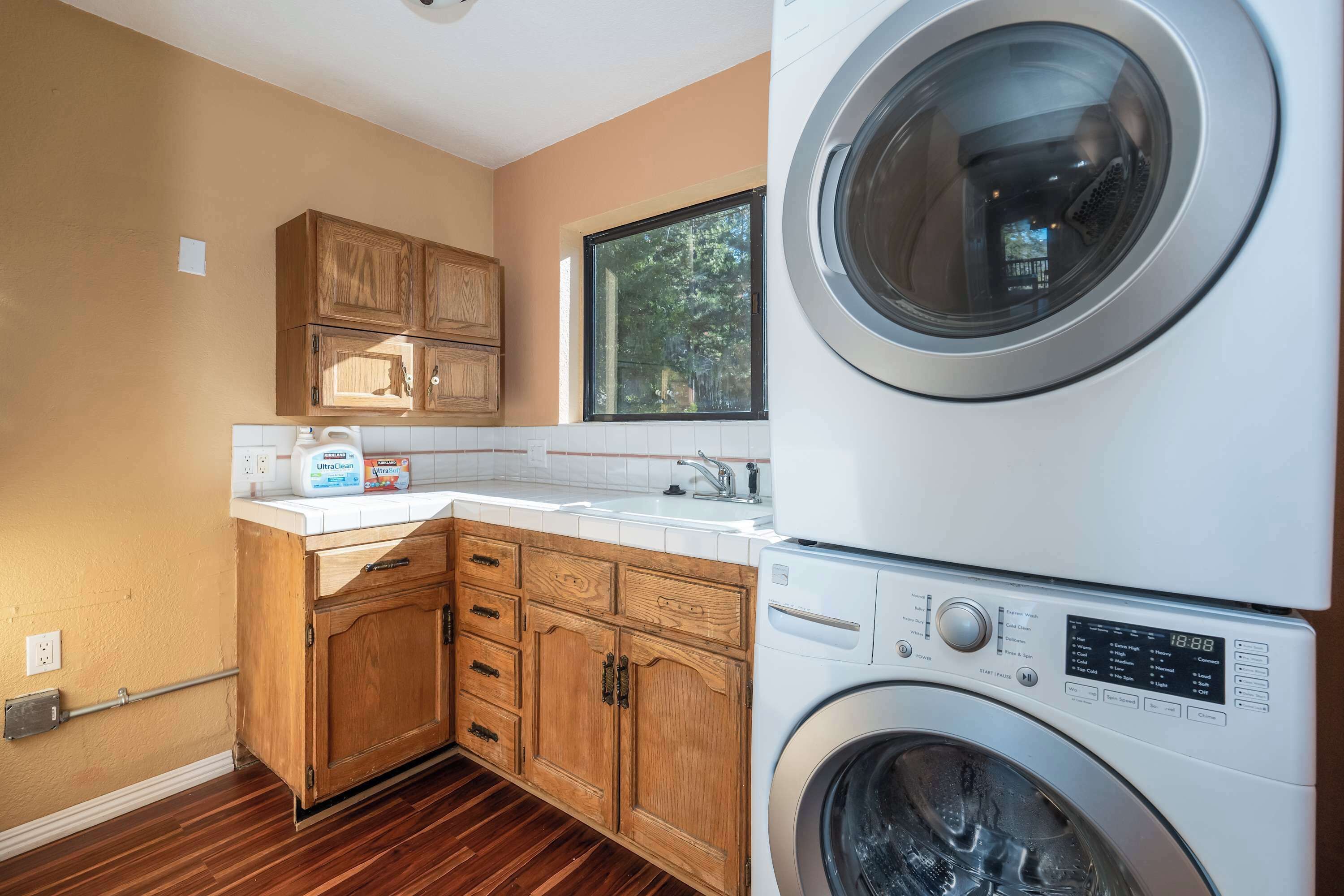 A laundry room with a stacked washer and dryer, wooden cabinets, a window, and a countertop with cleaning supplies.