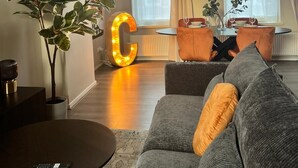 A modern living room with a large illuminated 'C', a grey sofa, a round coffee table, and a dining area with orange chairs and a small table. - Zwart Janstraat  (Rotterdam)