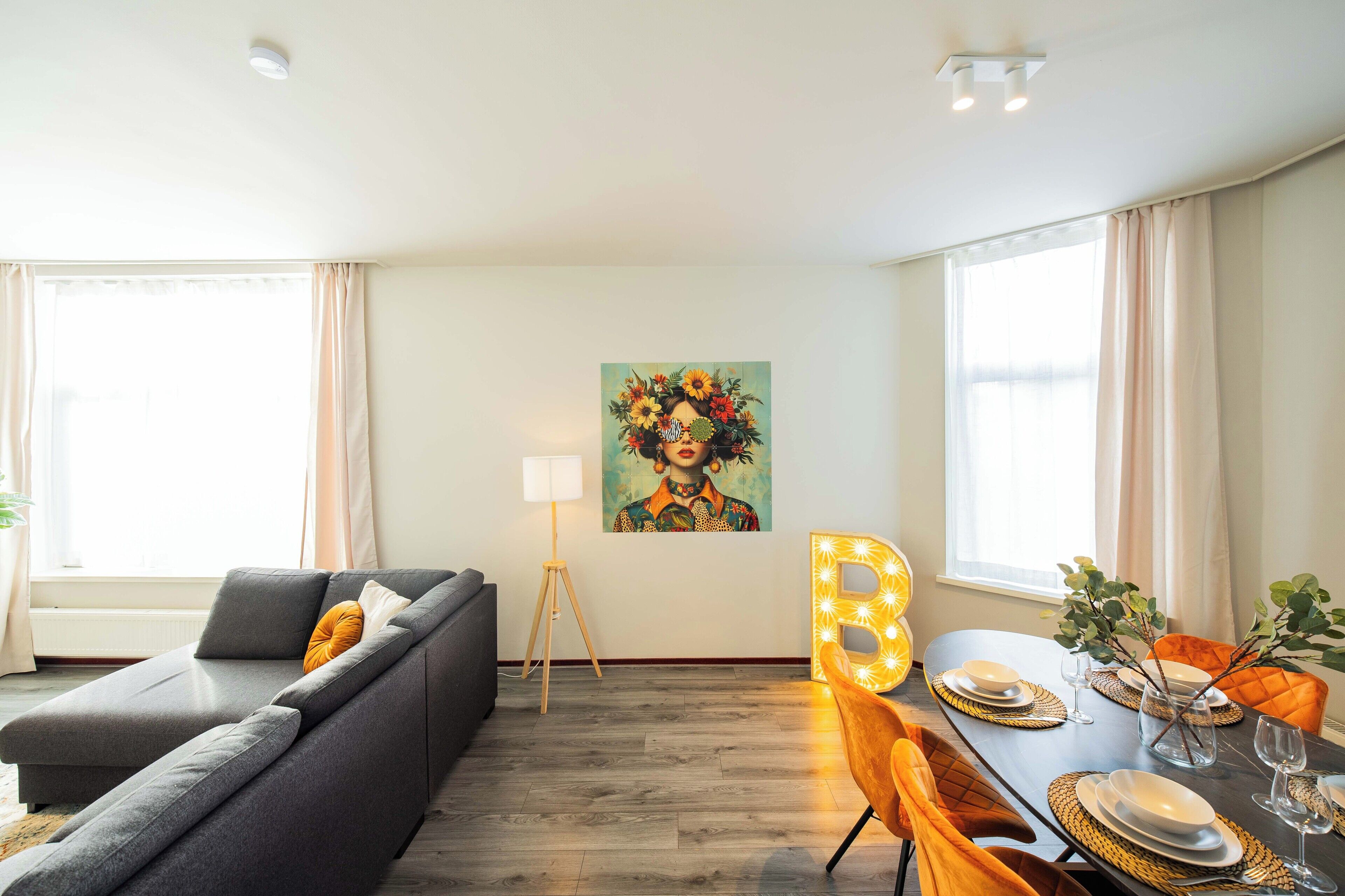 A modern living room with a grey sofa, a dining table set for four, and a large letter 'B' illuminated sign.