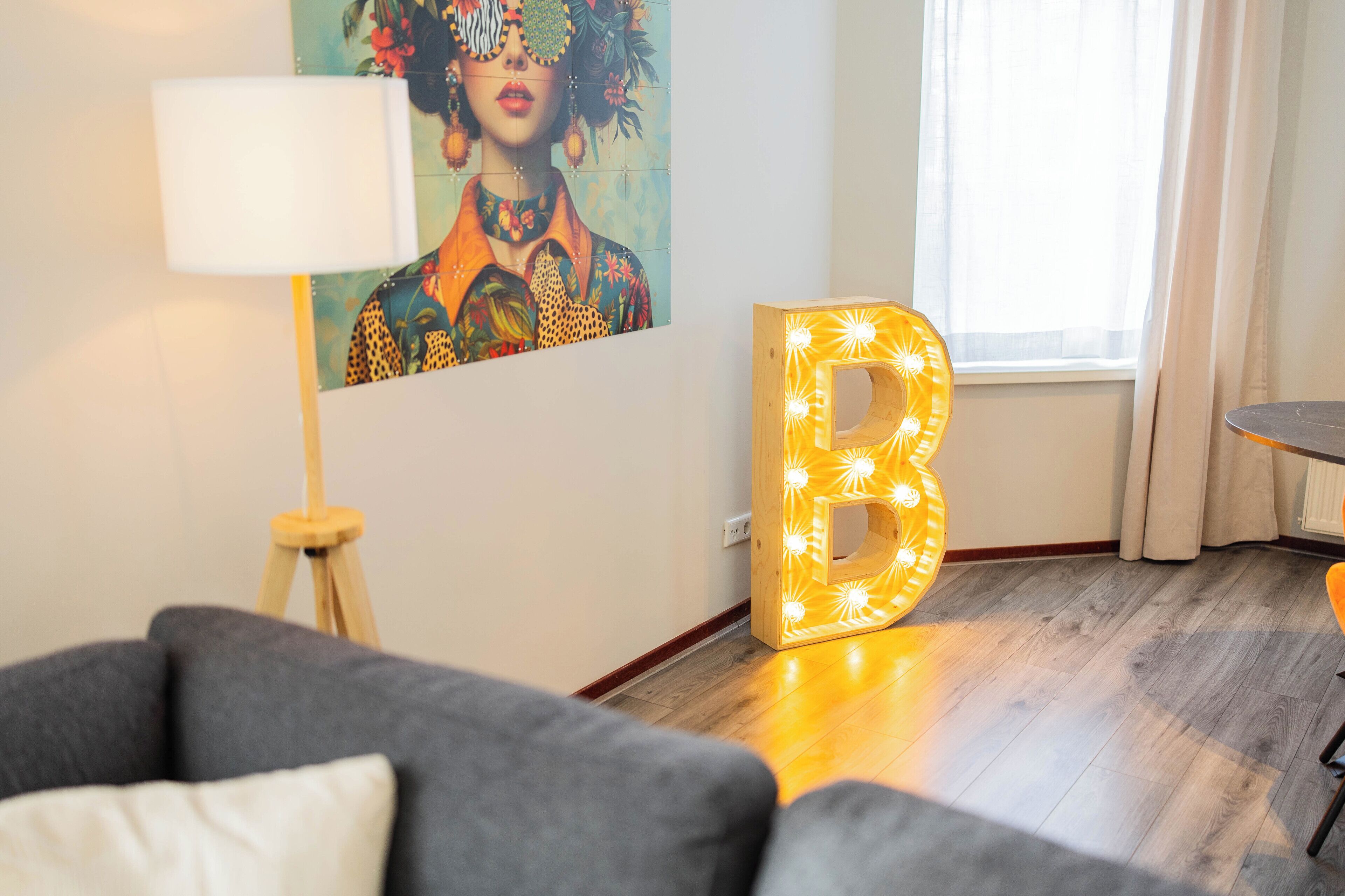 Een moderne woonkamer met een grijze bank, een lamp, een groot verlicht 'B' bord en een kleurrijk schilderij aan de muur.