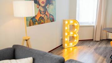 Een moderne woonkamer met een grijze bank, een lamp, een groot verlicht 'B' bord en een kleurrijk schilderij aan de muur.