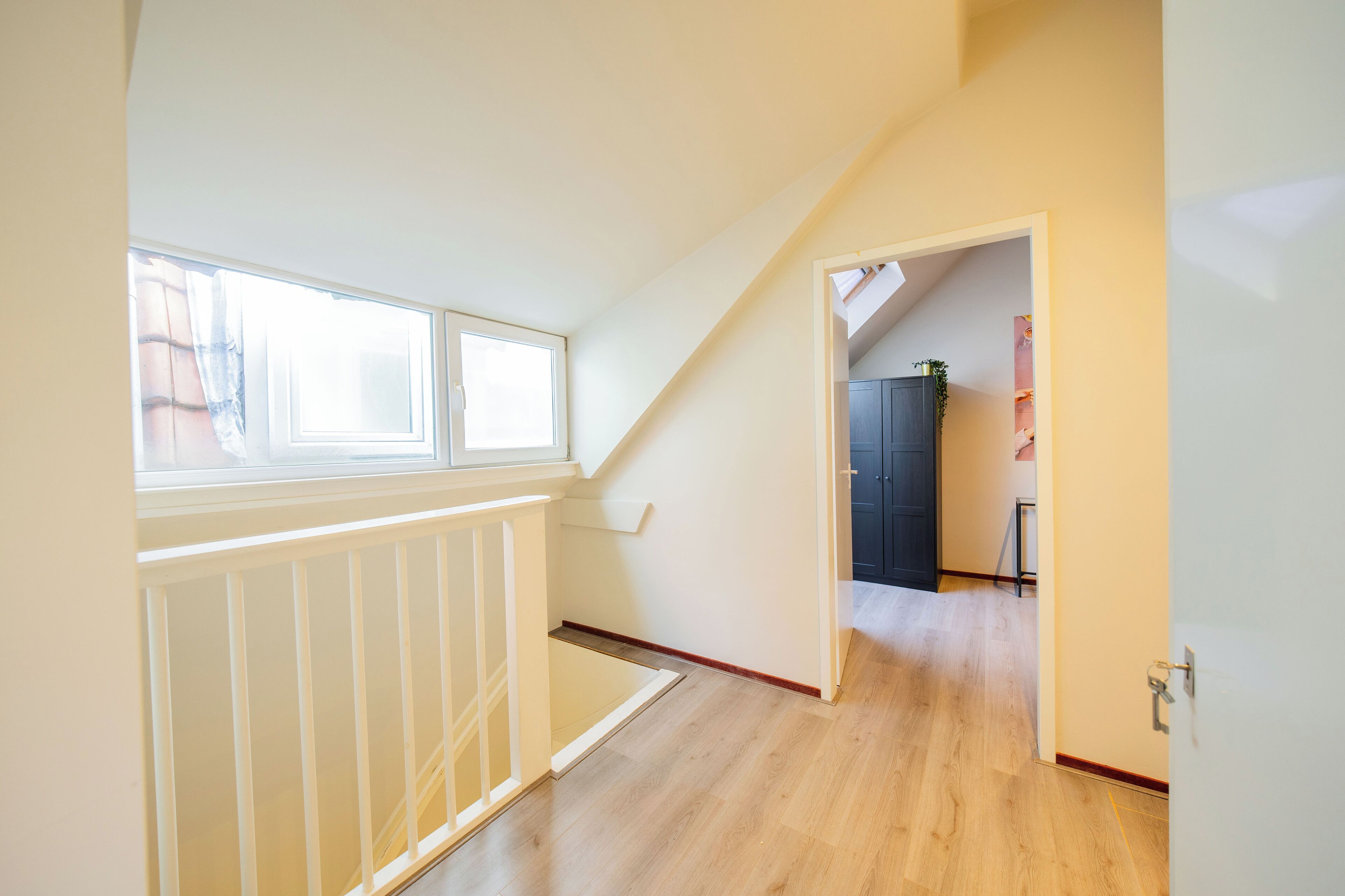 A narrow hallway with a wooden floor, a white railing, and a door leading to a room with a dark-colored cabinet and a small table with a plant.