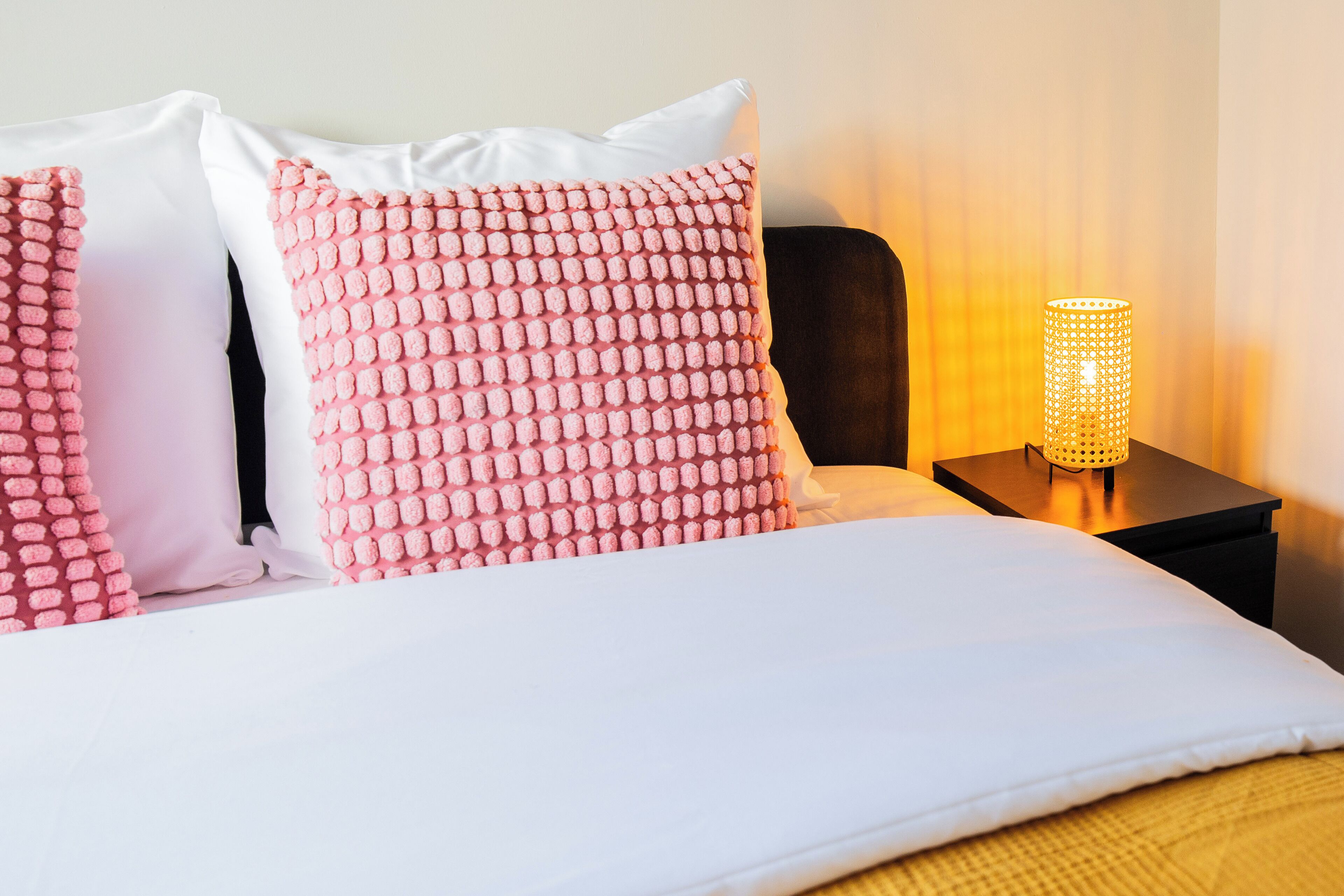 A neatly made bed with white and pink pillows, a bedside lamp, and a dark nightstand.