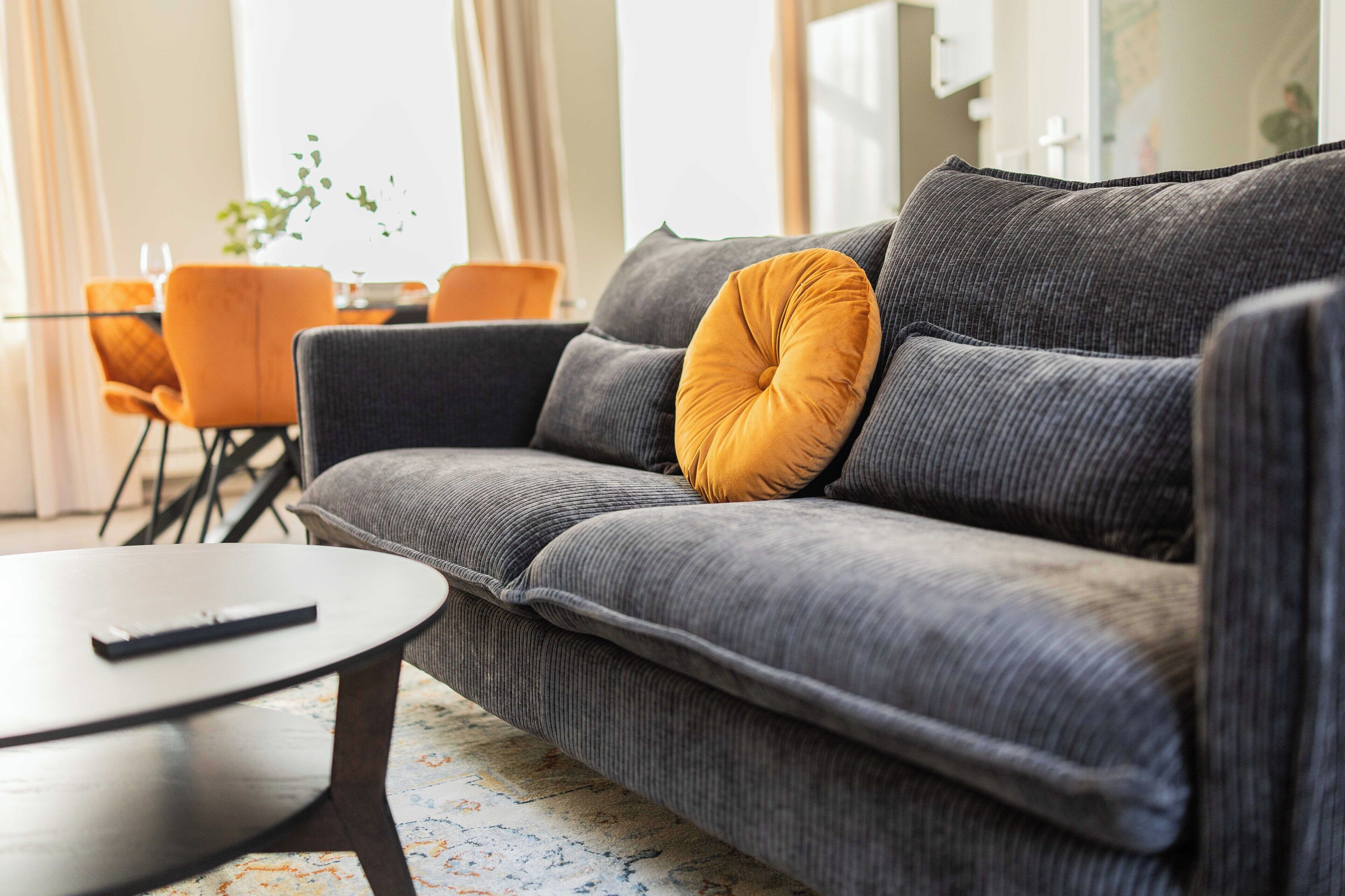 A grey sofa with a mustard yellow pillow, a remote control on a round coffee table, and orange chairs in the background.
