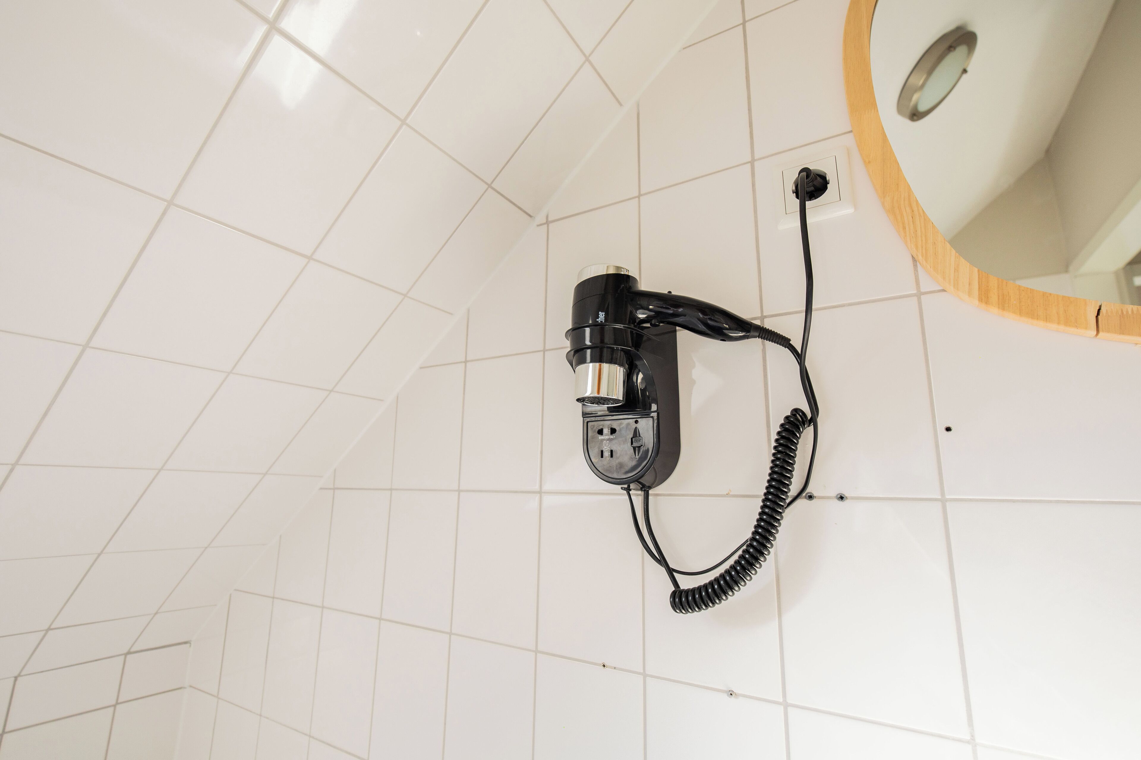 Une salle de bain avec un sol carrelé de carreaux blancs, un sèche-cheveux noir fixé au mur et un miroir encadré en bois.
