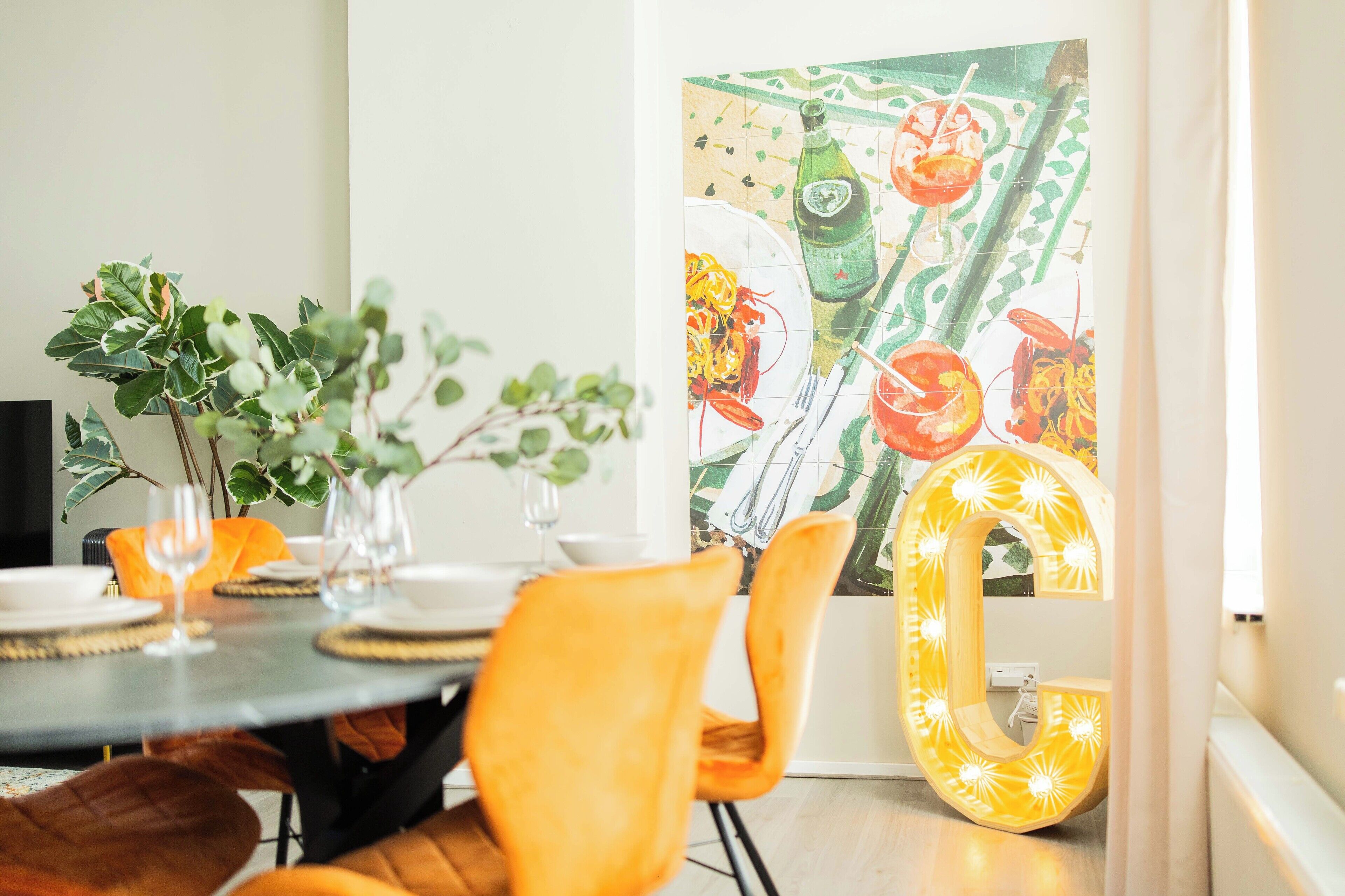 Een eetgedeelte met een ronde tafel voor vier personen, omringd door oranje stoelen. Achtergrond toont een grote, verlichte letter 'L'.