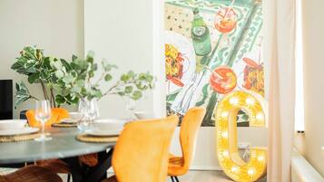 Een eetgedeelte met een ronde tafel voor vier personen, omringd door oranje stoelen. Achtergrond toont een grote, verlichte letter 'L'.