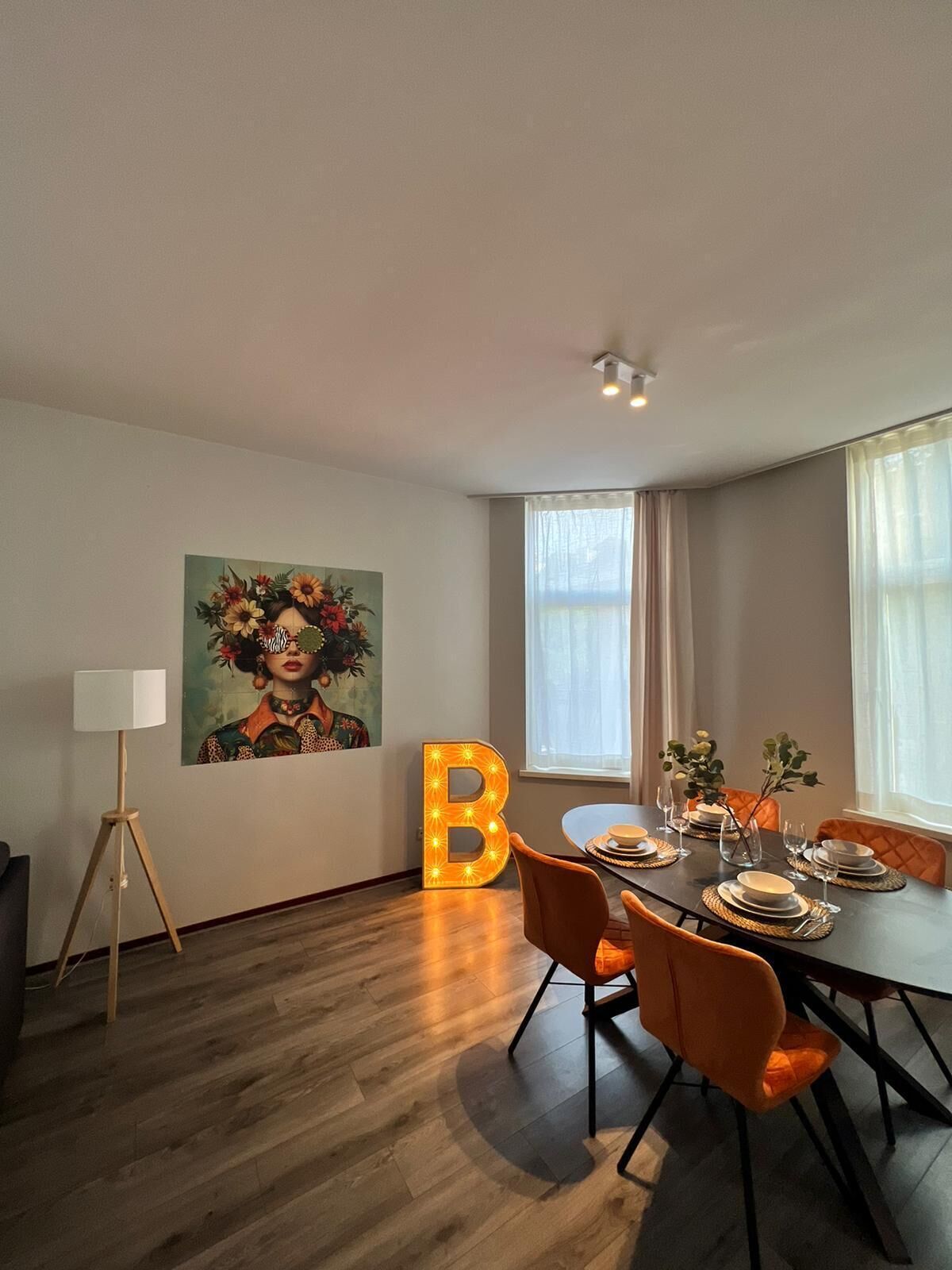 Un comedor con una mesa dispuesta para cuatro, una gran 'B' iluminada en la pared y un cuadro sobre la mesa.