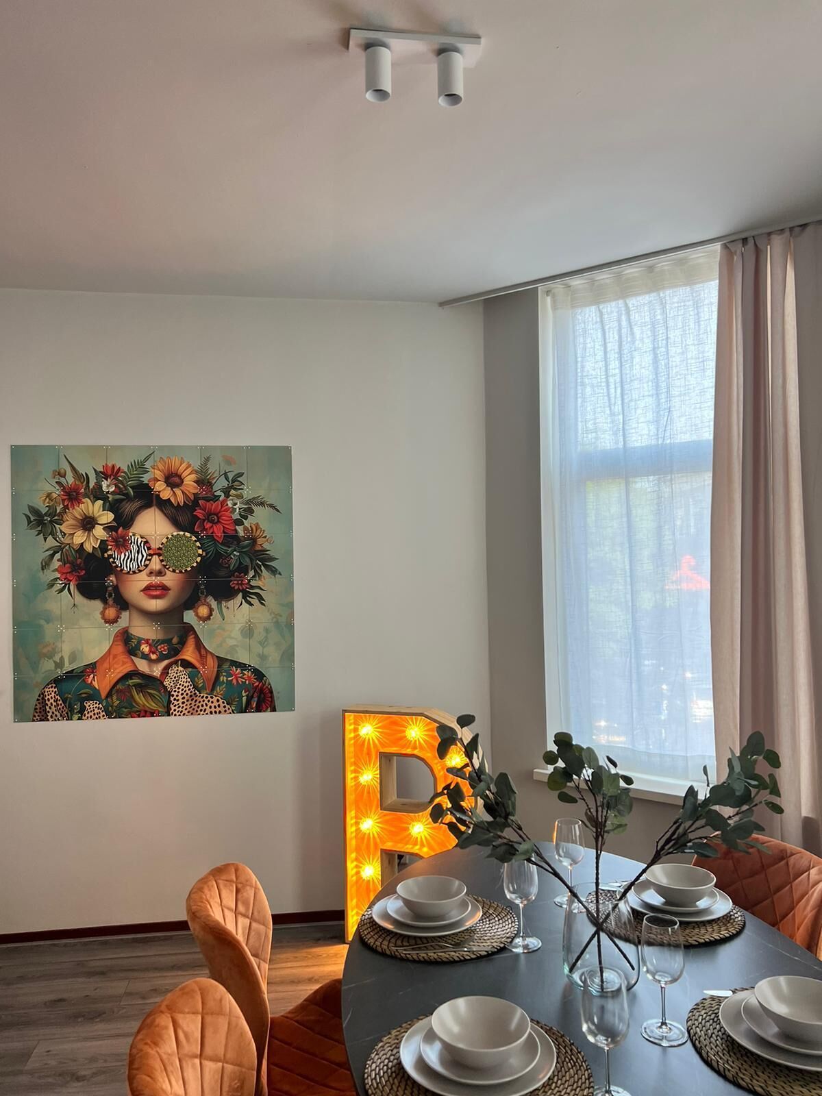A dining area with a table set for four, a large illuminated letter 'D', and a vibrant painting on the wall.