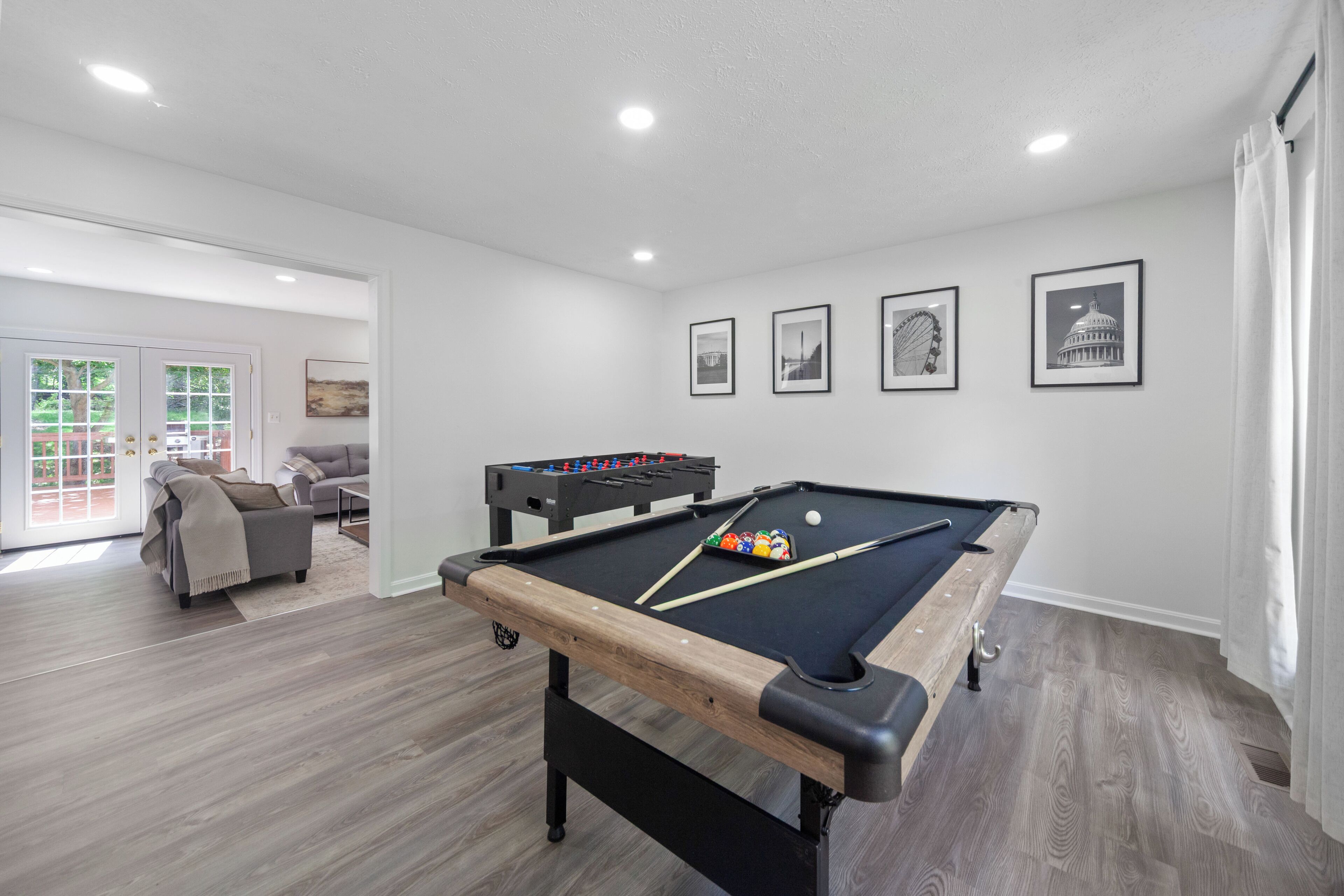 A pool table with billiard balls, a foosball table, and a sofa in a room with wooden flooring and white walls.