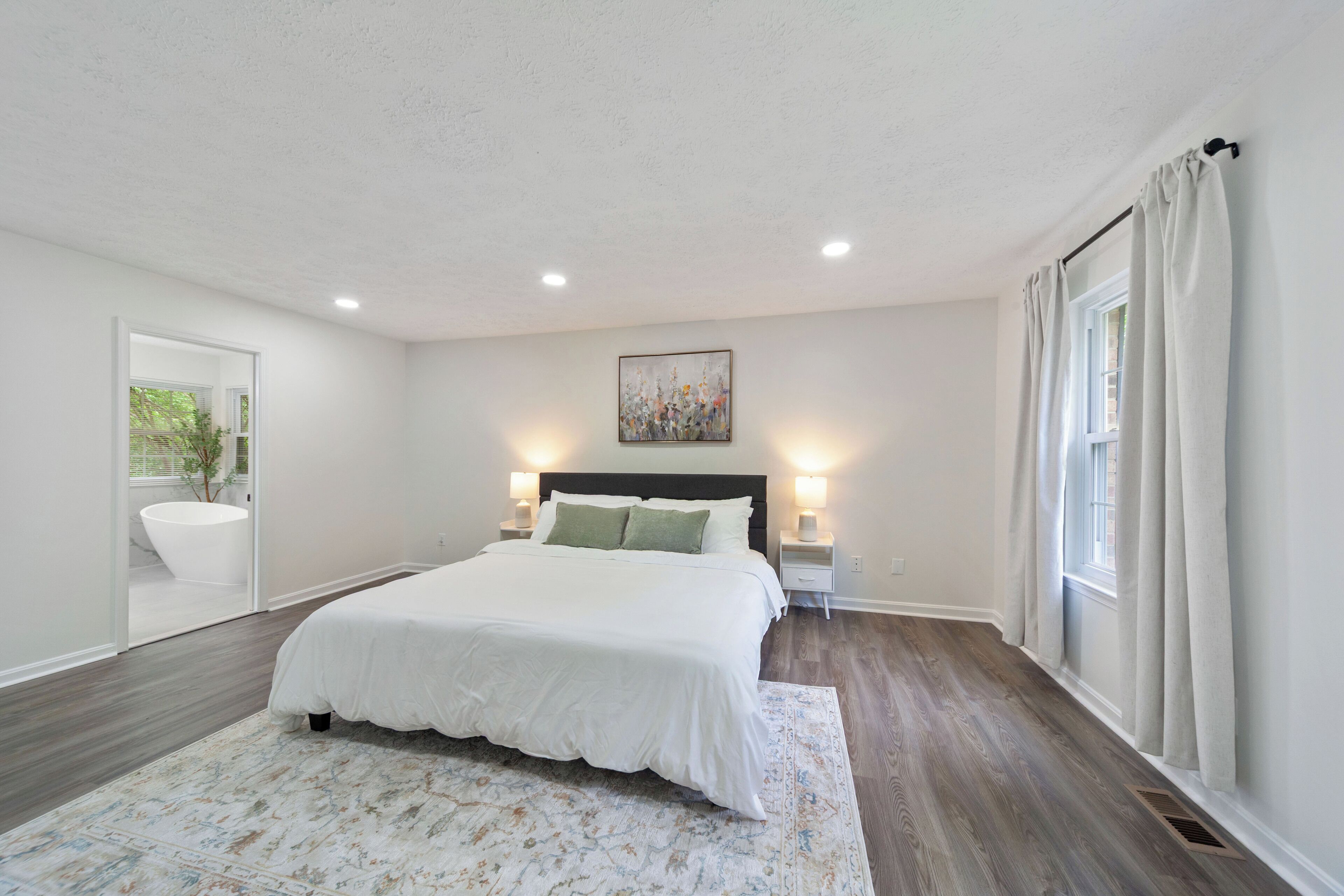 A bedroom with a large bed, bedside lamps, a painting on the wall, and a view of a bathroom through an open door.