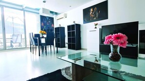 A modern living room with a glass coffee table, a dining area with chairs, a TV, and artwork on the walls.
