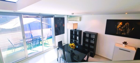 Un salon moderne doté d’une porte vitrée donnant sur un balcon, d’un coin repas avec une table et des chaises, et d’une étagère noire.