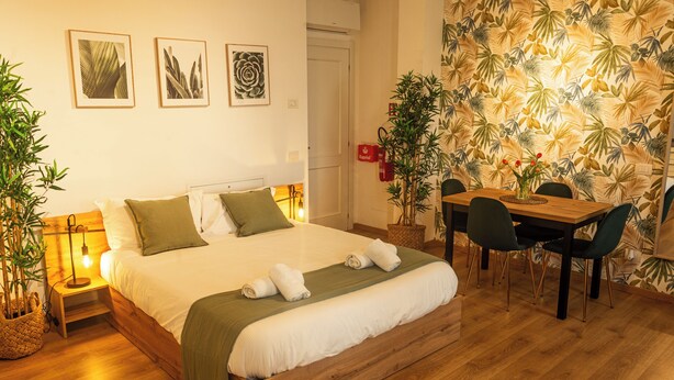 A bedroom with a bed, bedside table, lamp, and a dining area with chairs and a table.