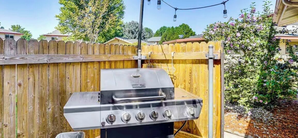 Una parrilla de acero inoxidable con cuatro quemadores, conexión de gas y panel de control, frente a una valla de madera y arbustos florecientes.