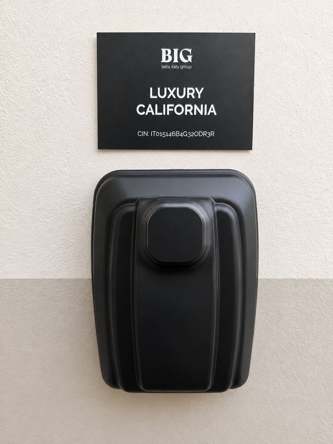 A black suitcase with a handle, mounted on a wall above a black card with 'BIG beta italy group LUXURY CALIFORNIA' text.