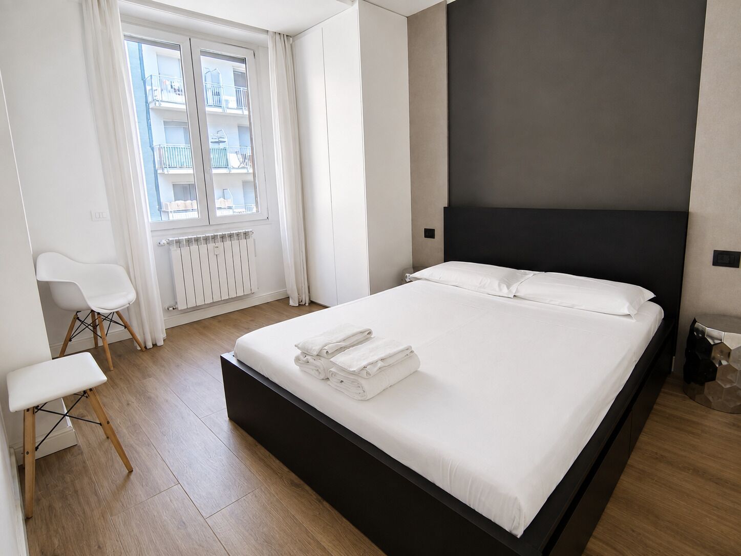 Ein modernes Hotelzimmer mit einem großen Bett, einem weißen Stuhl, einer Heizung und einem Fenster mit Blick auf andere Gebäude.