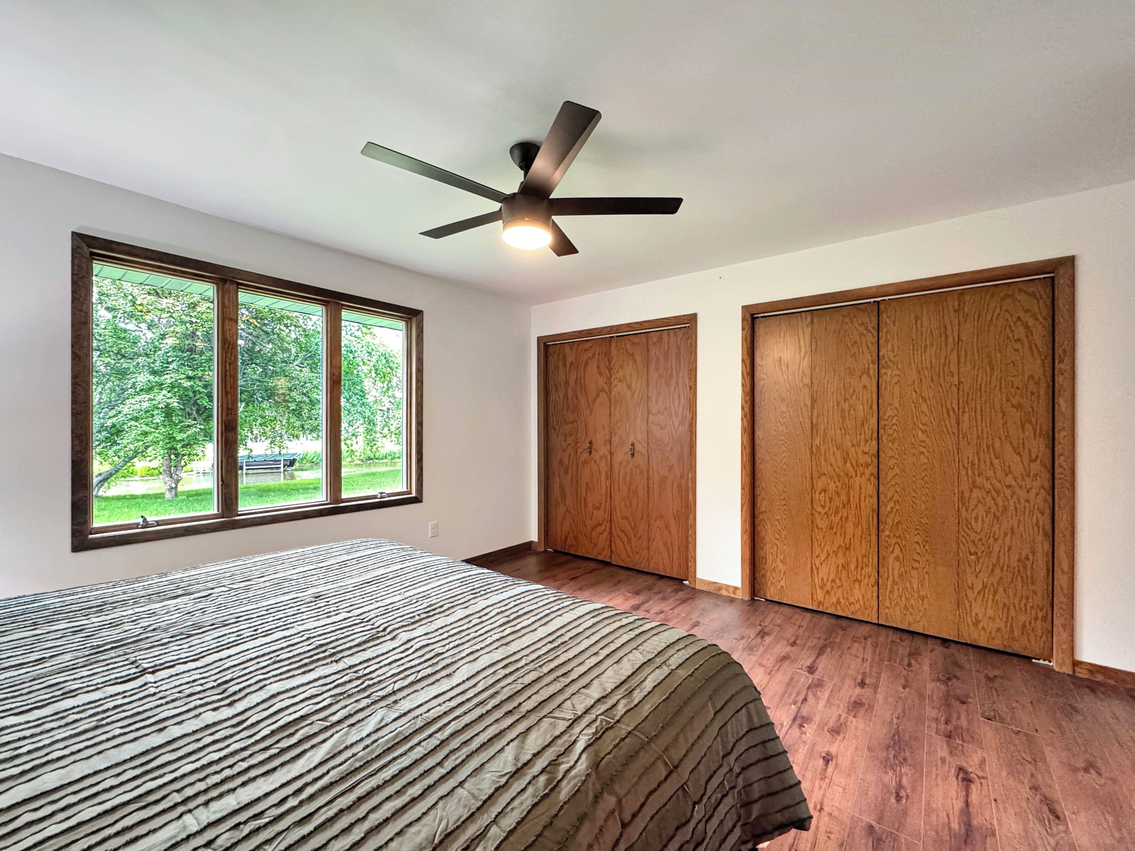 Une chambre à coucher avec un lit, un ventilateur de plafond, une grande fenêtre et des armoires en bois.