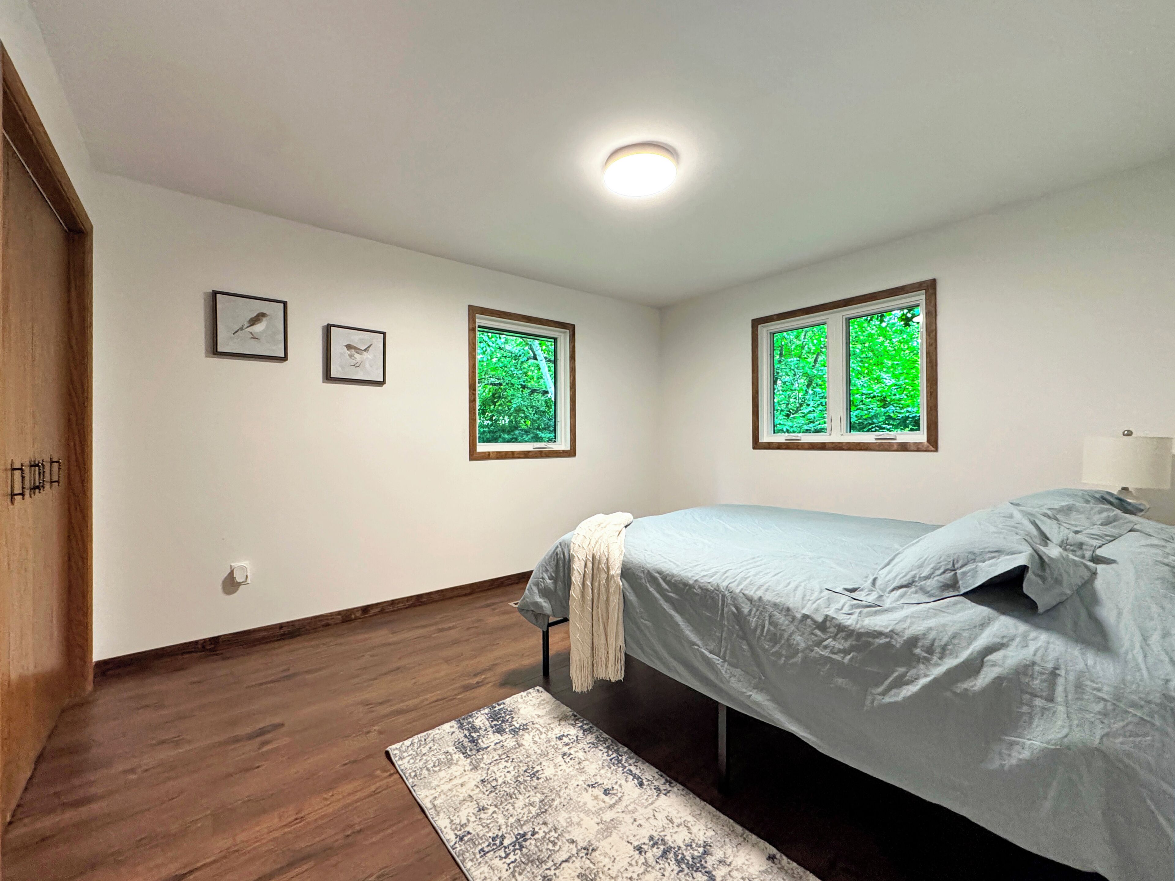Un dormitorio con una cama, dos ventanas que dan a follaje exterior, piso de madera y una puerta.