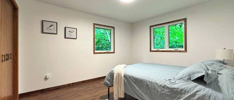 Un dormitorio con una cama, dos ventanas que dan a follaje exterior, piso de madera y una puerta.