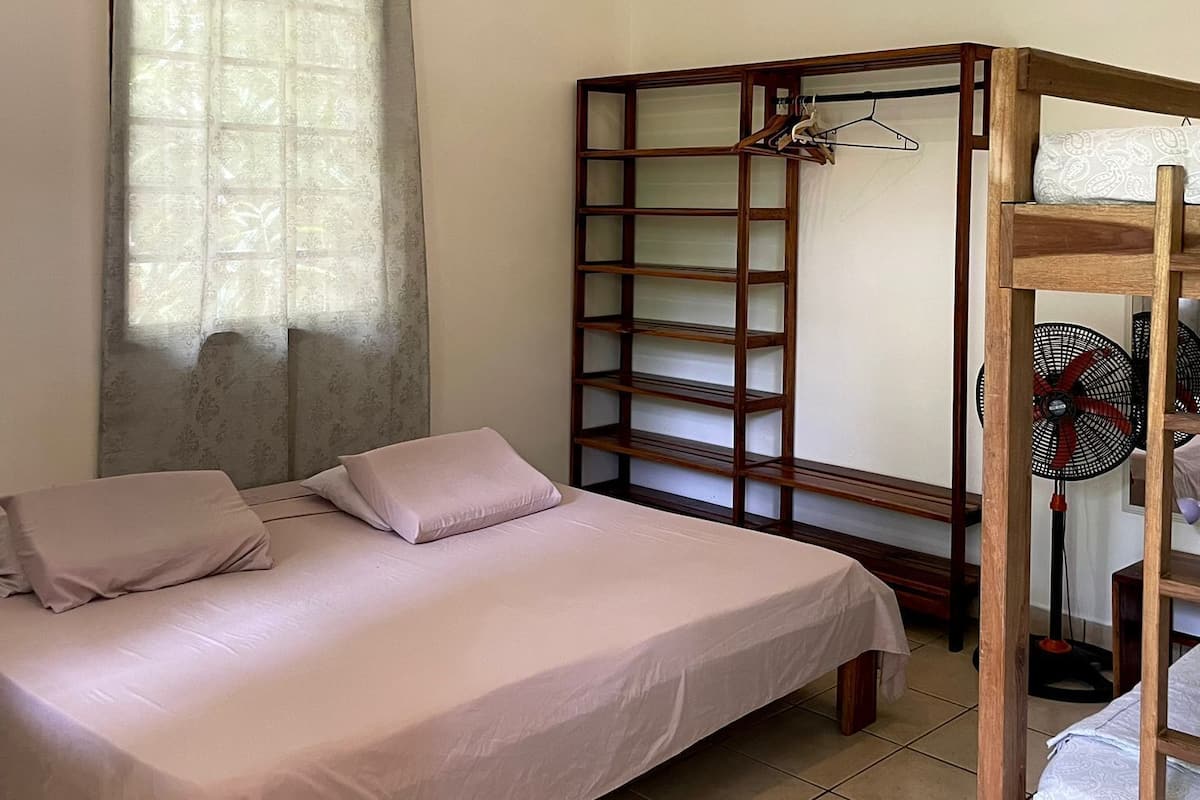 Quarto com cama, guarda-roupa de madeira, ventoinha e janela com cortinas.
