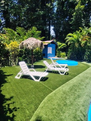 A pool area with artificial grass, two white lounge chairs, a thatched-roof structure, and a small blue building.