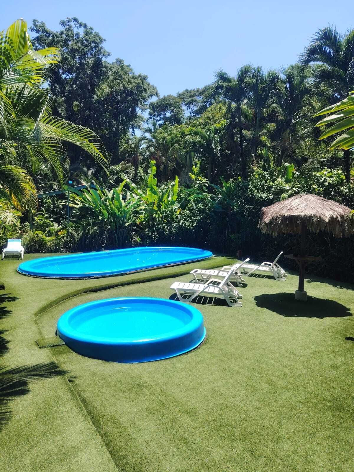 A backyard with two swimming pools surrounded by artificial grass, a thatched umbrella, and lounge chairs.