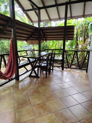Un patio techado al aire libre con una mesa y sillas, rodeado por una barandilla y abundante vegetación.