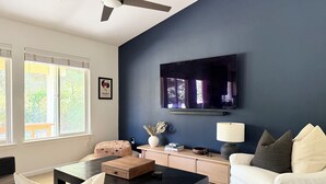 Un salon avec un mur d’accent bleu foncé, un téléviseur à écran plat, un ventilateur de plafond, un canapé blanc, une table basse noire et une fenêtre avec des stores.