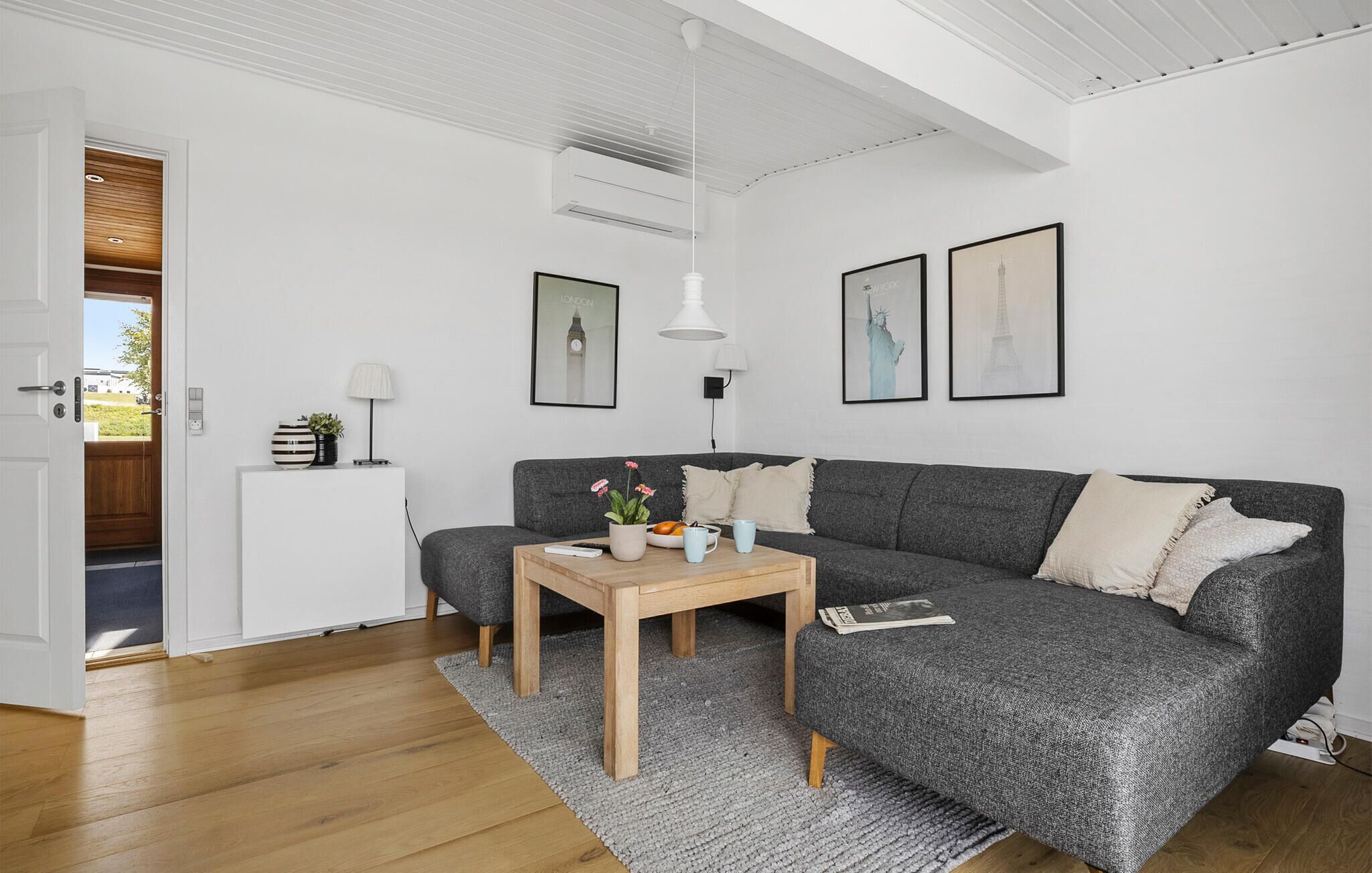 Una sala moderna con un sofá seccional gris, una mesa de centro de madera y una alfombra gris. Hay dos cuadros enmarcados en la pared, una lámpara y una pequeña mesita con una planta.