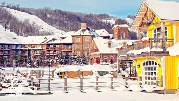 Un village enneigé, avec des bâtiments traditionnels en bois, un imposant bâtiment jaune doté d’un balcon, et une piste de ski en arrière-plan.