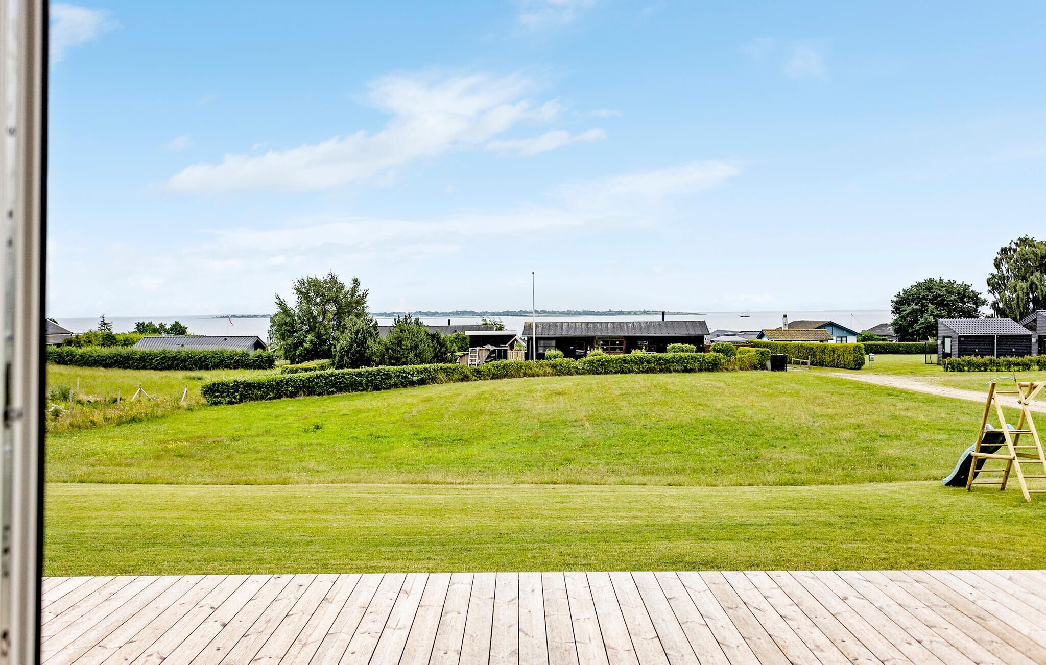 A view of a grassy area with a wooden deck in the foreground, a body of water, and several buildings in the background.