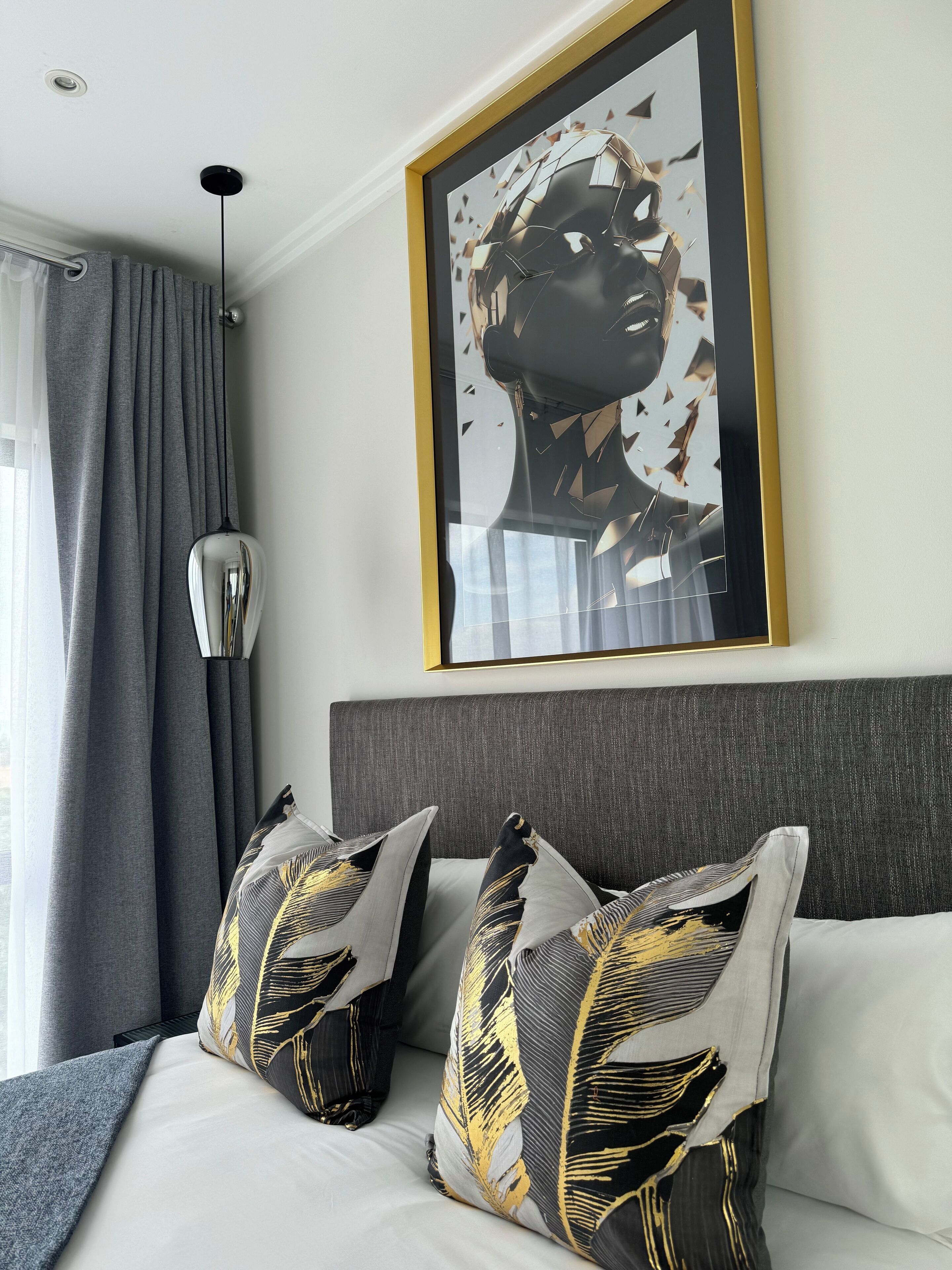 A modern hotel room with a large bed, grey curtains, a framed artwork on the wall, and a unique pendant light.