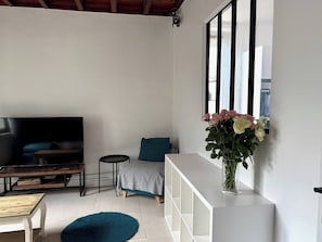 Una sala moderna con un televisor de pantalla plana, un sofá, una mesita redonda pequeña, un tapete azul y un florero sobre una consola blanca.
