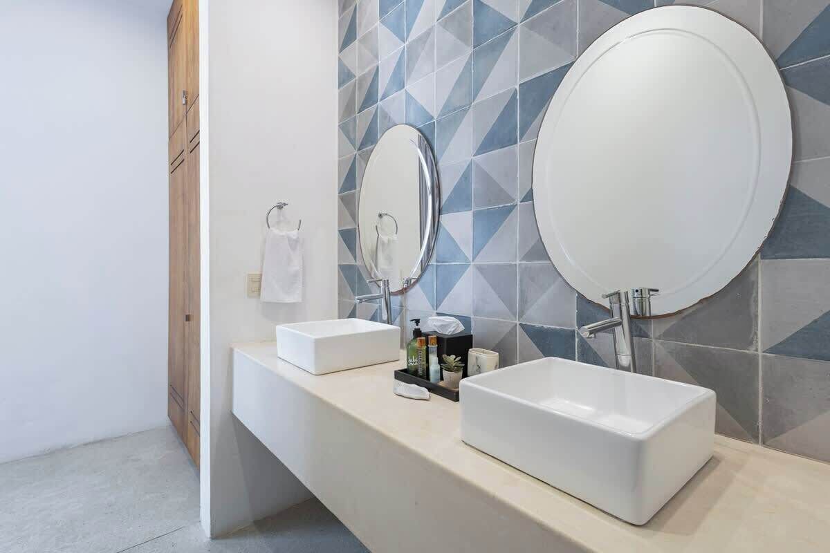 Une salle de bain moderne avec deux lavabos blancs, un miroir et un mur recouvert de carreaux.
