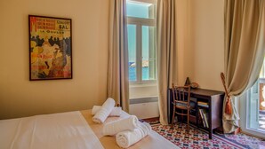 A hotel room with a bed, two rolled towels, a desk with a chair, a poster on the wall, and a window with curtains. - A charming retreat with timeless beauty (Andros Chora)