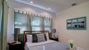 Un dormitorio ordenado con cama, mesitas de noche, una ventana con cortinas, un cuadro en la pared y una bandeja con flores.
