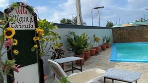 Zona junto a la piscina con un letrero que dice "Villa Mamá Carmita", rodeada de plantas en macetas y mobiliario de exterior.