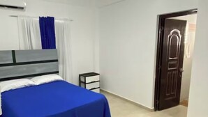 Un dormitorio con una cama, un ventilador de techo, una mesita de noche pequeña y una puerta.