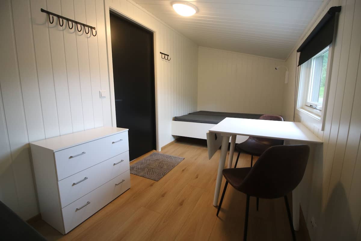 Een kleine, praktische kamer met een bed, een bureau, een stoel en een kastje.