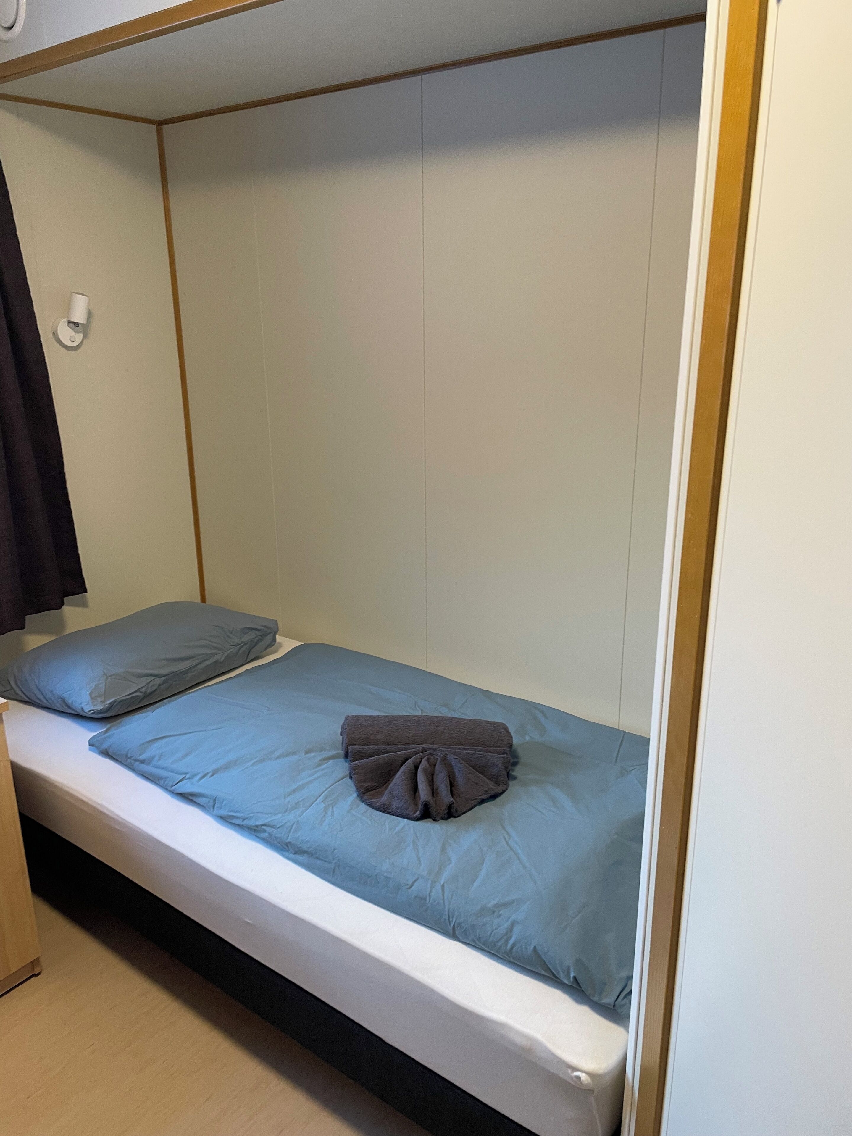 A single bed with a blue mattress and pillows, a wooden headboard, and a white wall with a light switch.