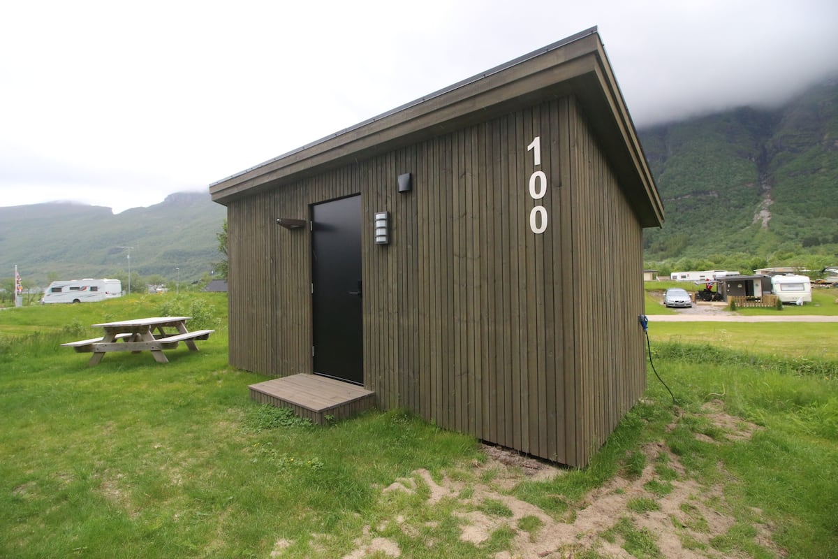 Een klein houten vakantiehuisje met een deur, een bankje en het nummer '100' aan de zijkant.