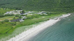 Vue aérienne d’une zone côtière comprenant une plage, un petit village avec des maisons et une route, ainsi qu’une forêt dense en arrière-plan.