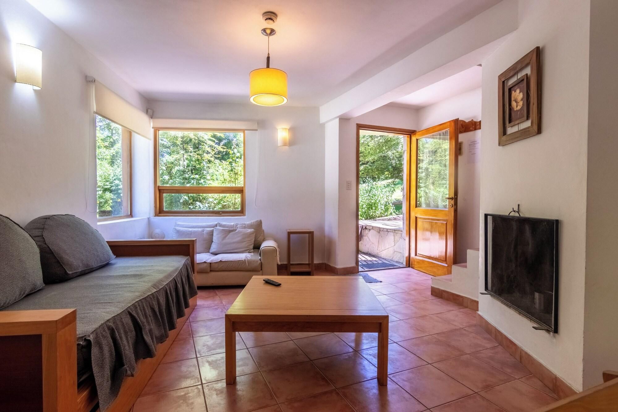 A cozy living room with a sofa, a coffee table, a TV, and a large window offering a view of greenery.